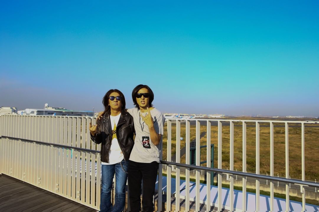 B'z公式SNSで公開された「足湯スカイデッキ」で肩を組む松本孝弘と稲葉浩志の写真